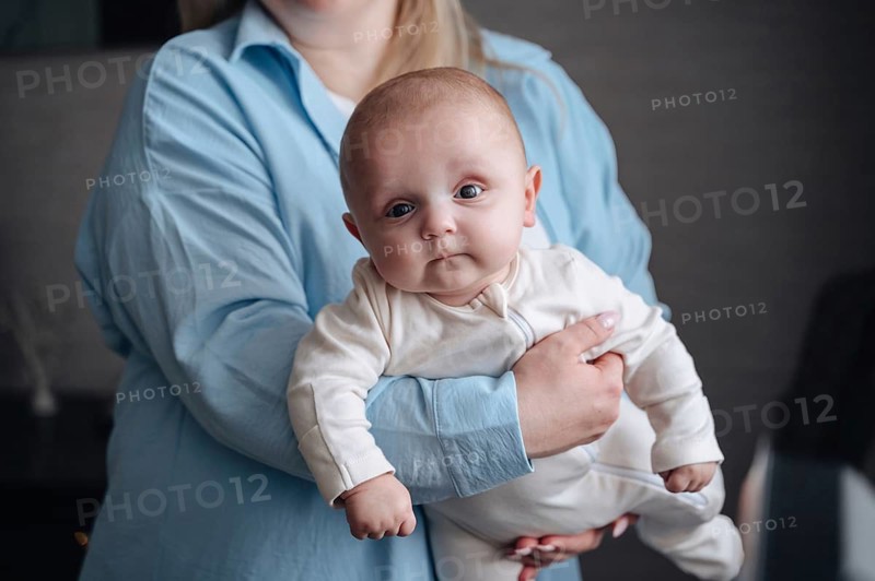 A cute baby boy gazes curiously while being held gently by his mother. The intimate setting conveys warmth and tenderness, highlighting the bond between parent and child
