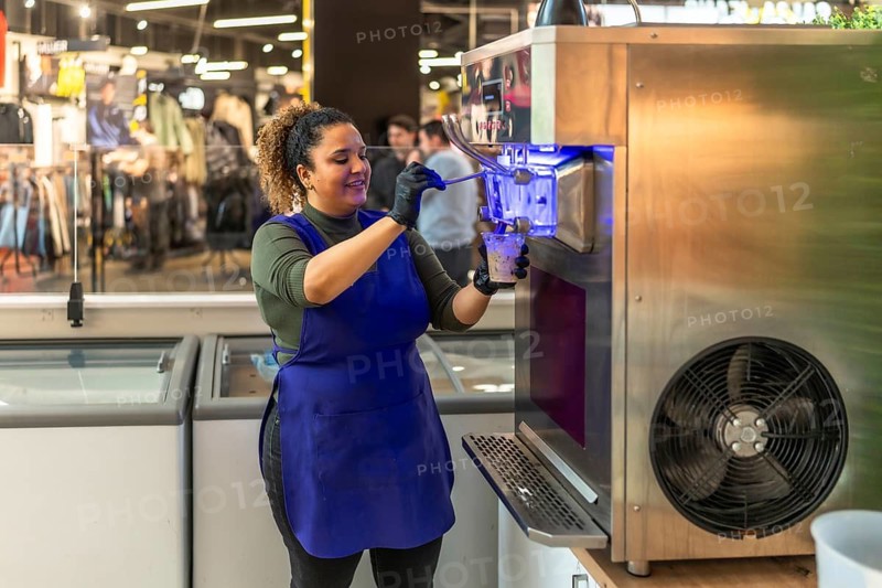 Hispanic woman in blue apron and black gloves smiling while serving soft serve ice cream from a commercial machine into a plastic cup at a shop counter, indoor daytime scene