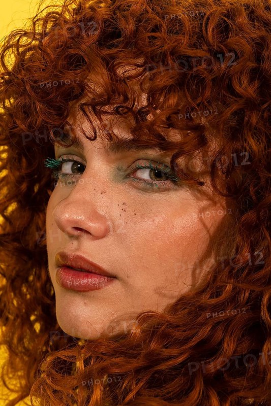 A striking portrait of a woman with curly red hair. Her expressive eyes, accentuated by bold makeup, create a captivating and vivid look against a bright background