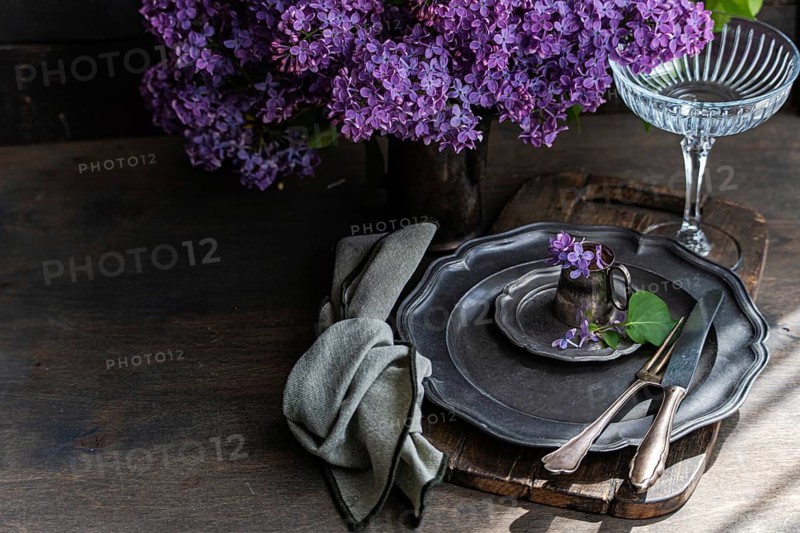 Elegant table setting with vintage plates, silverware, and delicate lilac flowers on a rustic wooden board. A soft, textured cloth napkin completes the charming arrangement