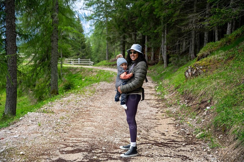 Mother carries her baby in her arms during a walk in a lush forest at the foot of the Dolomites. The path winds through tall trees and green meadows, capturing a peaceful and happy moment in nature