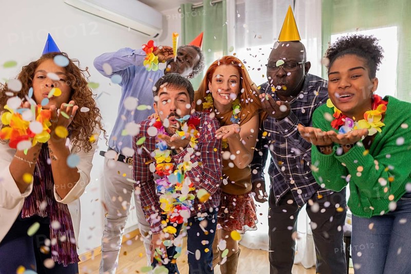 A lively group of friends celebrating a birthday indoors, blowing colorful confetti, wearing party hats and leis. The room is filled with laughter and festive joy