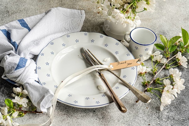 A beautifully arranged table setting features a white plate with blue patterns, a fork and knife tied with a ribbon, a Thank You tag, and white flowers adding elegance