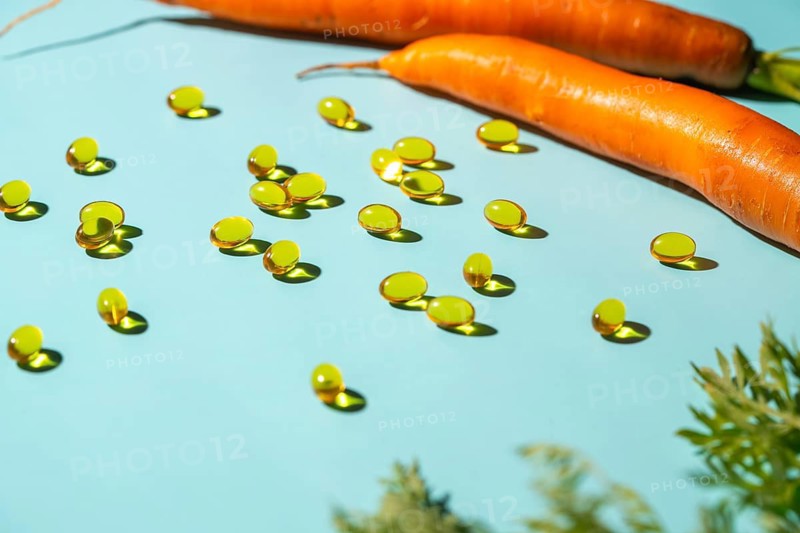 A vibrant image of softgel vitamin capsules spread across a light blue surface alongside fresh carrots, symbolizing health and wellness through natural supplements