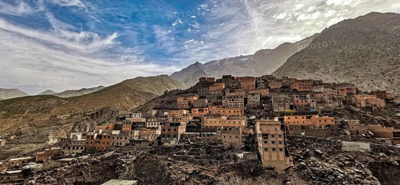 Dramatic shot of a mountain village with clay-colored houses nestled against a range of hills in atmospheric colors under a sunny sky with white clouds, Morocco