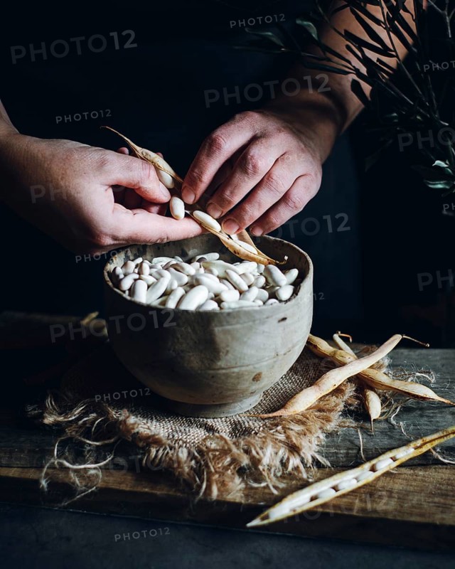 Close-up of hands shelling fresh white beans into a rustic wooden bowl, surrounded by empty pods on a textured cloth. A tranquil scene capturing the essence of home cooking