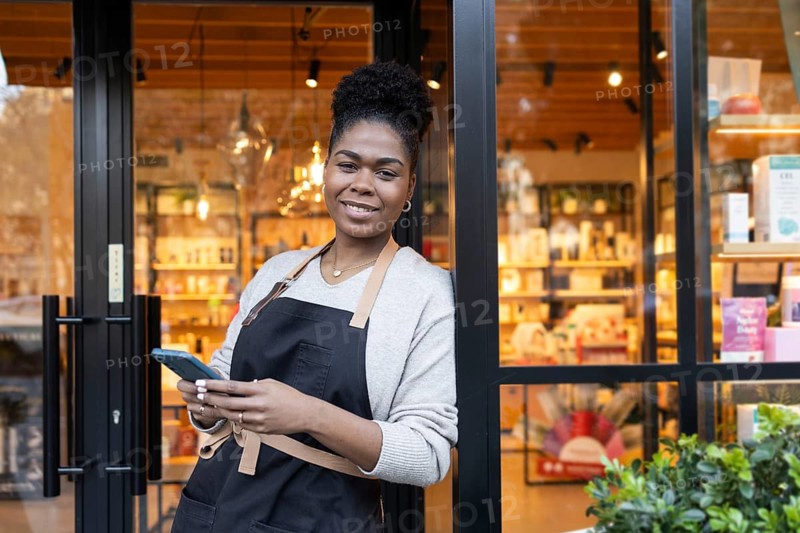 Smiling worker in apron stands outside a vibrant cosmetics store, holding a smartphone. Shelves with colorful products are visible through the large glass window
