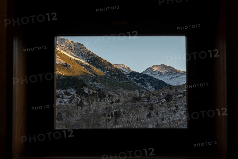 A serene winter scene showcasing snow-capped mountains viewed through a dark, framed cabin window, highlighting nature's tranquility and the season's beauty