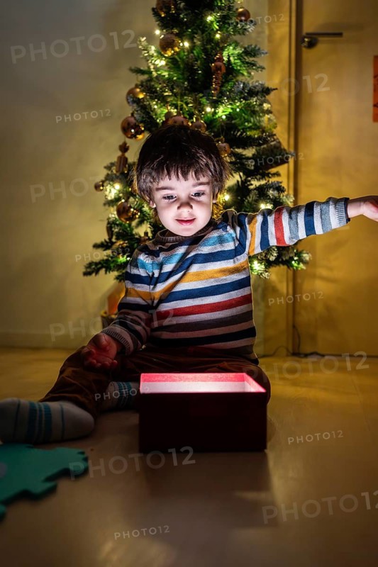A young boy is sitting in front of a Christmas tree and opening a red box. Scene is joyful and festive, as the boy is surrounded by the holiday season