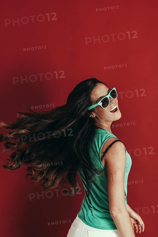 A woman with long hair, wearing green sunglasses and a teal top, smiles and turns her head playfully against a vibrant red background, exuding joy and energy