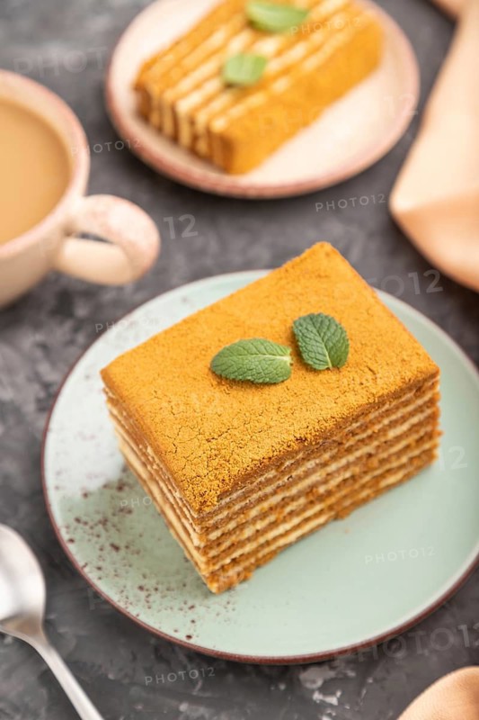 Homemade honey cake with milk cream and mint with cup of coffee on a black concrete background and orange textile. Side view, close up, selective focus