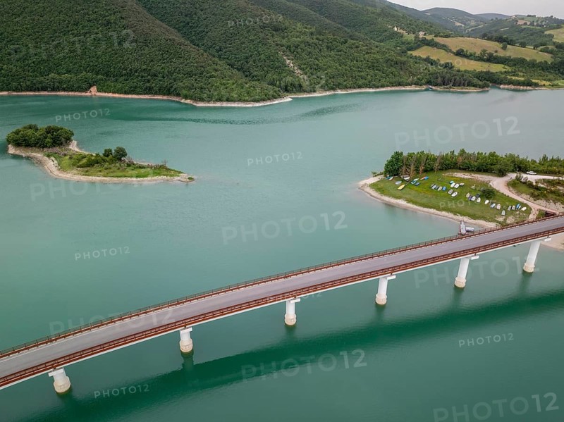 A serene aerial view of a bridge stretching across the calm waters of Castreccioni Lake in Italy. Surrounded by lush greenery, the scene captures the essence of peaceful travel