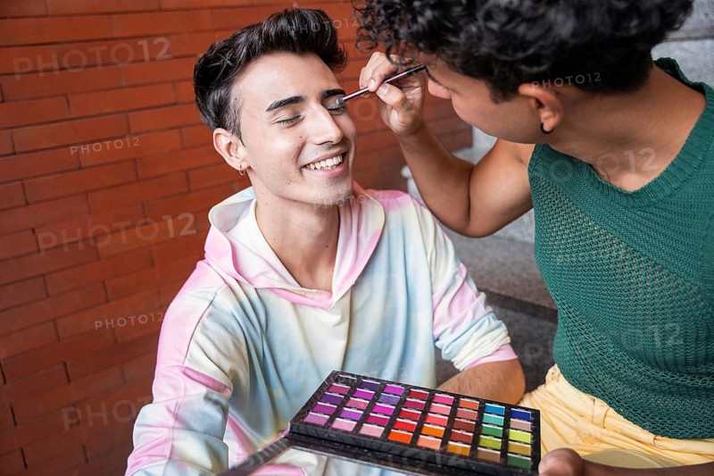 A happy couple enjoys a makeup session outdoors. One person applies colorful eyeshadow on the other's eyelid, creating a joyful and intimate moment on a sunny day