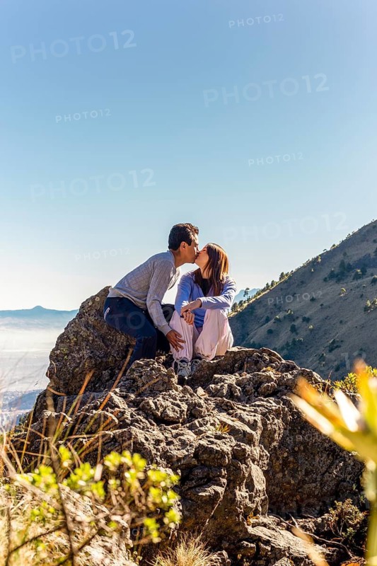 A couple shares a heartfelt kiss while sitting on top of a rocky mountain The sun is shining brightly, creating a warm and cozy atmosphere under a clear blue sky
