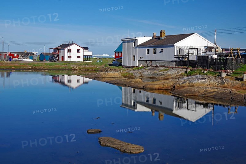 Simple houses are reflected in a calm lake surrounded by rocks and clear blue skies