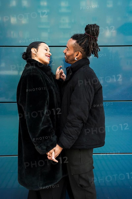 A multiethnic couple stands outdoors in cozy jackets, sharing a joyful moment as they smile warmly at each other. A celebration of diversity and love