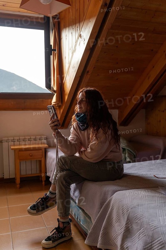 A redheaded woman sits comfortably on a bed in a wooden cabin, holding a smartphone and looking at screen, with a serene mountain view visible through the window