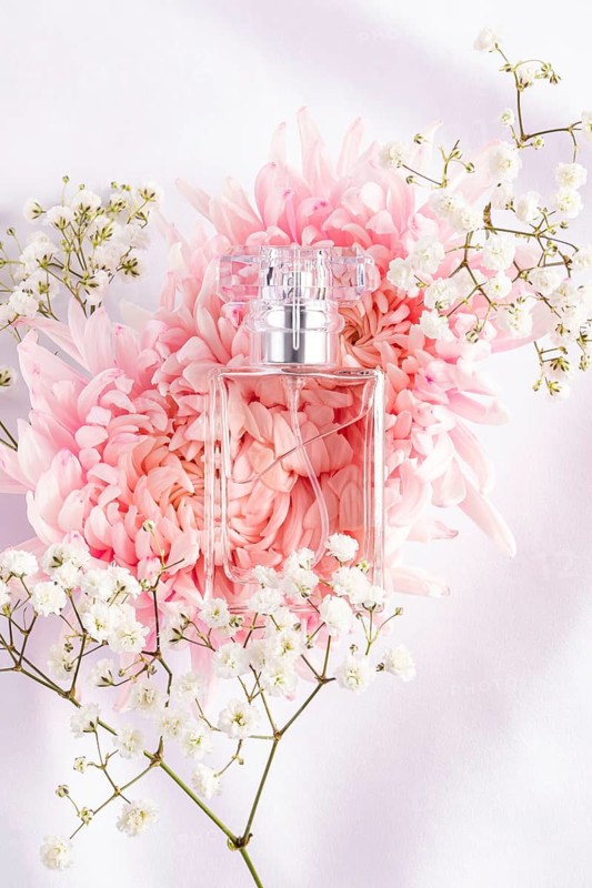 A transparent glass perfume bottle is elegantly placed on a background of pink chrysanthemums and delicate white baby breath flowers, creating a fresh and sophisticated aesthetic