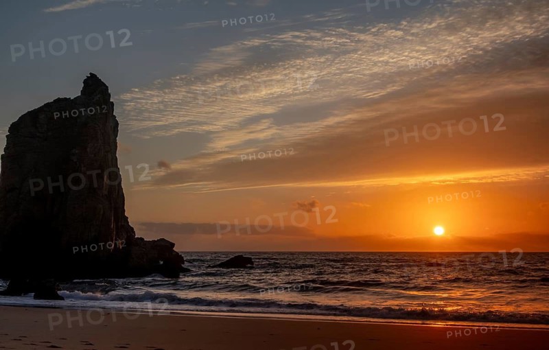 Scenic view of a towering rock formation at sunset, with the sun casting a warm glow over the ocean waves and sandy beach, creating a tranquil, picturesque scene in bear beach, Portugal