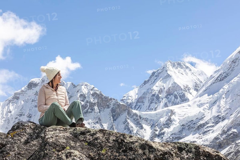 Woman sits on a rock, dressed warmly, gazing at the majestic snow-capped peaks of the Everest region. Clear blue sky and rugged landscape enhance the breathtaking scenery