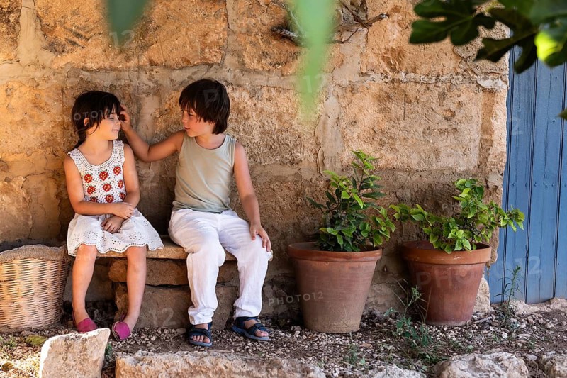 Two children sit on a stone bench by a rustic wall, engaging in conversation. They are surrounded by potted plants, creating a warm, inviting scene