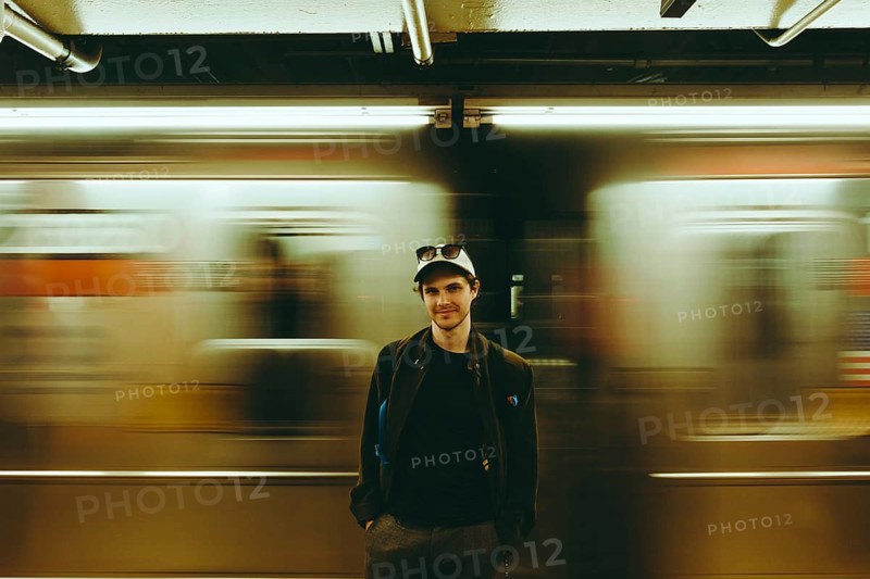 A man wearing a cap and jacket stands in a subway station with a blurred train moving in the background in New York. The image captures the essence of urban travel and movement