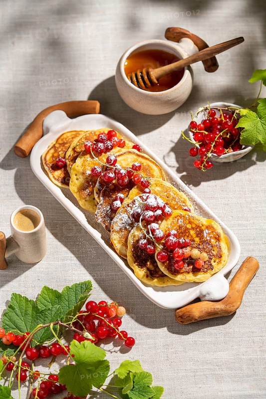 Top view of delicious pancakes topped with red currants on a white ceramic tray. The light linen background enhances the freshness, with honey and coffee alongside