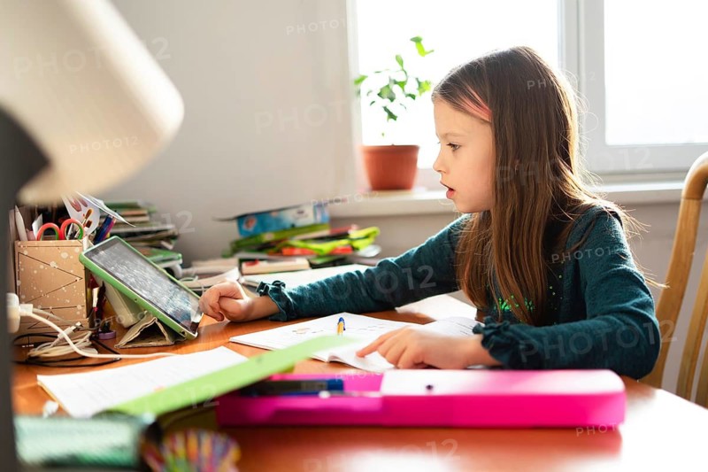A young schoolgirl sits at her desk, focused on her homework. The scene includes a tablet and school supplies, illustrating a typical study session at home