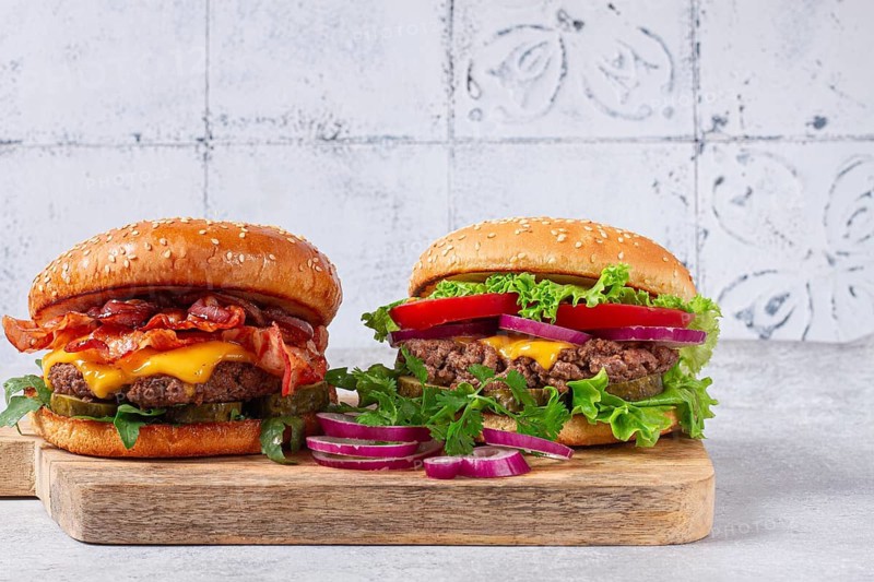 Two burgers with beef patty, fried bacon, cheddar cheese, tomatoes, on a light table