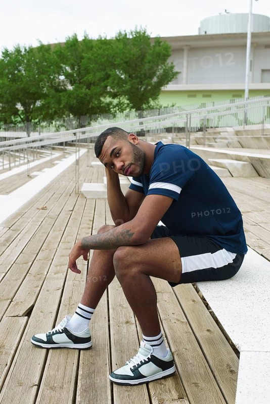 A black male athlete in sporty attire sits on wooden steps, taking a break from training. He appears reflective, surrounded by a modern urban backdrop
