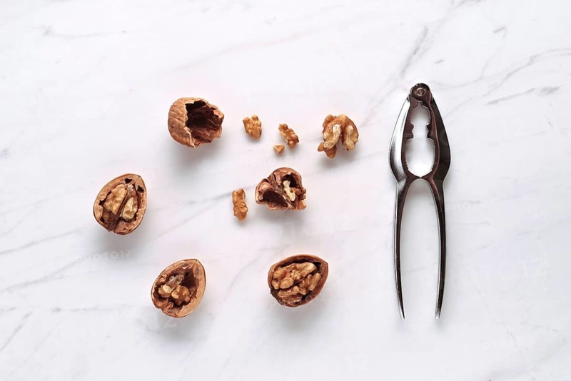 Cracked walnuts and a metal nutcracker on a white marble countertop. The arrangement highlights the rustic texture of the walnuts and the sleek design of the tool