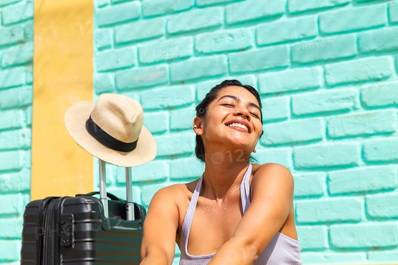 A young Latin woman smiles brightly, enjoying a sunny day. She sits beside a black suitcase and a straw hat in front of a colorful wall
