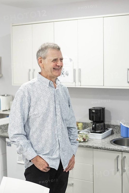 A man with intellectual disabilities stands smiling in a bright, modern kitchen with light cabinets. The atmosphere is relaxed and welcoming, capturing a peaceful moment at supervised home