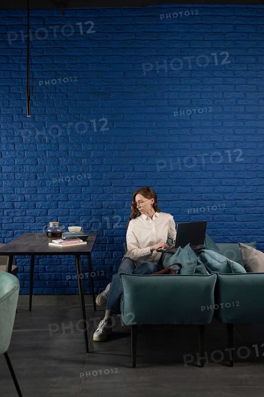 Woman in casual attire sitting on a modern couch with a laptop. Blue brick wall background, books, and tea on a table. Cozy and stylish interior ambiance