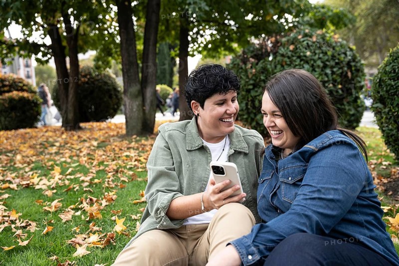 A joyful lesbian couple laughs together while looking at a phone, seated in a park with fallen autumn leaves and lush greenery around them on a peaceful day