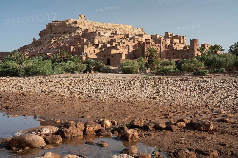 A stunning view of an ancient Moroccan kasbah, nestled in the rocky desert terrain Ksar of Ait Ben Haddou. The traditional architecture is surrounded by lush greenery, set against a clear blue sky