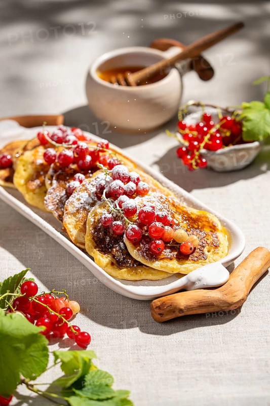 From above, a platter of golden pancakes topped with vibrant red currants sits on a light linen surface. A honey pot and fresh currants add to the inviting breakfast scene