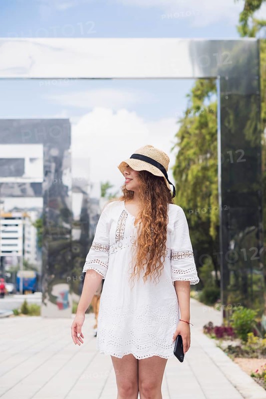 A young American Latin woman with long curly hair and a straw hat enjoys a sunny day outdoors. She wears a white dress and holds a phone, embodying modern urban lifestyle