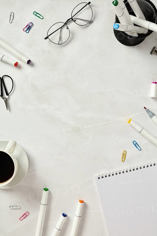 Top view of a creative and organized workspace featuring sketchbook, colorful markers, scissors, paper clips, and a coffee cup on a marble table. Includes glasses for a stylish look