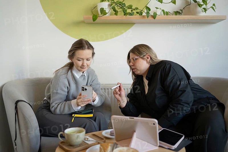 Two women engaging in conversation over coffee in a modern cafe setting. They are discussing work, with a smartphone and tablet on the table, surrounded by cozy decor