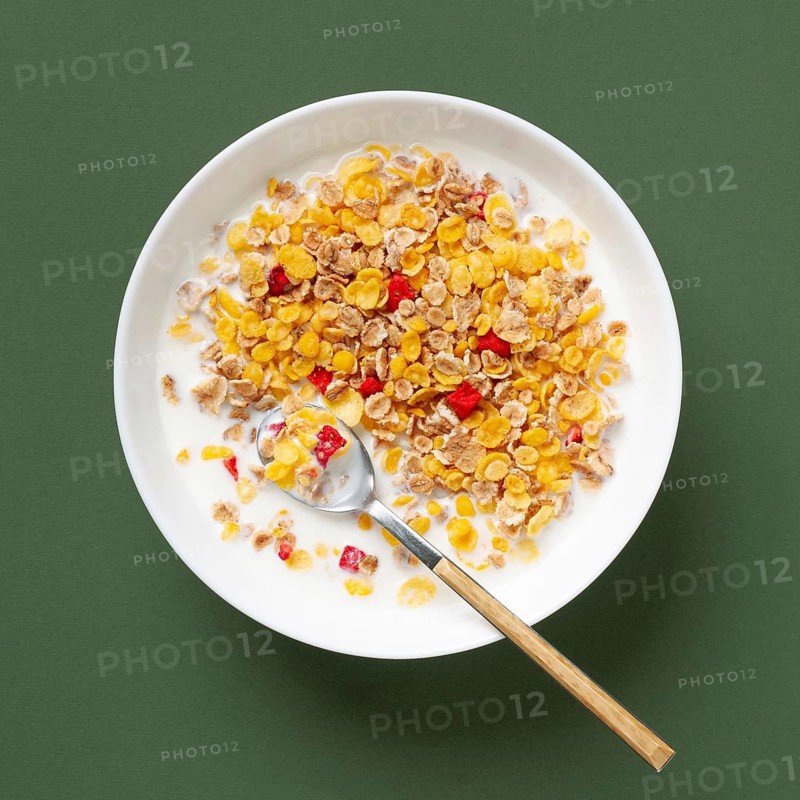 Top view of a nutritious bowl of cereals mixed with milk and colorful fruits on a green background. A wooden spoon rests in the bowl