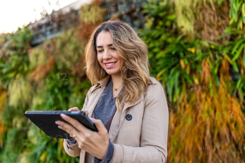 Woman smiling and wearing a trench coat while operating a digital tablet with her finger, standing outdoors against a vibrant vertical garden or living wall, embracing technology and nature