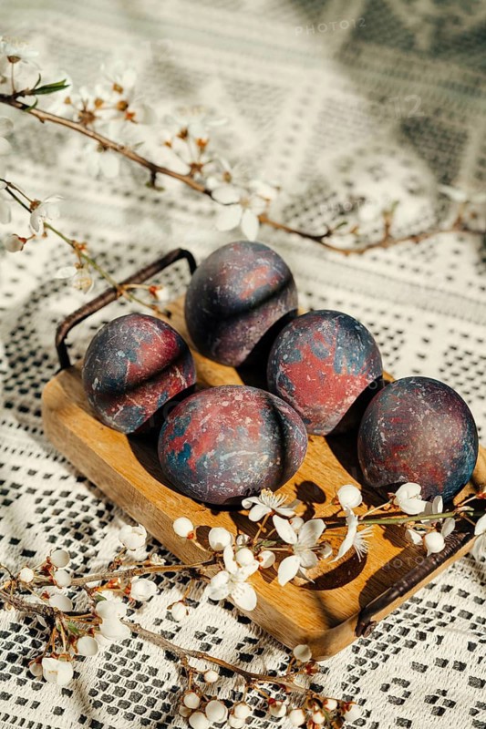 Artistic Easter eggs with a unique galaxy pattern displayed on a wooden tray, adorned with blooming spring branches, set on a crochet tablecloth textile
