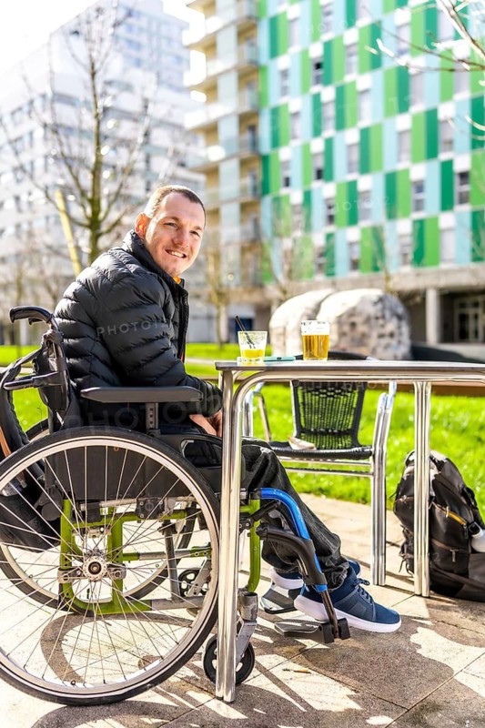 Young man in a wheelchair enjoys a refreshing drink with a friend at an outdoor cafe in the city, embracing urban life and social connection