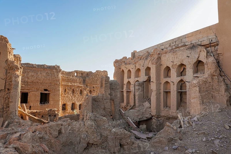 Weathered and crumbling structures in Al Hamra, an abandoned clay village, against a clear blue sky in Oman. This image captures the decay and historical ambiance of the site