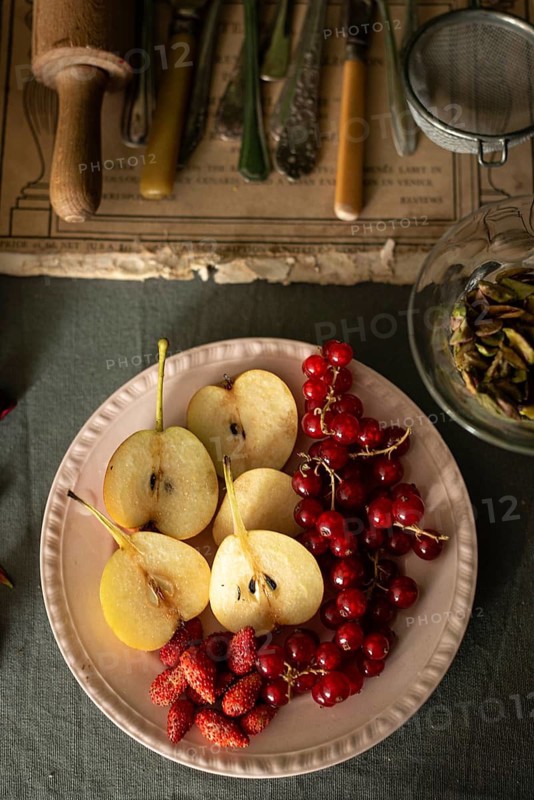 A vintage style setting features sliced pears, red currants, and strawberries on a plate. Rustic utensils are seen in the background, enhancing the autumn ambiance