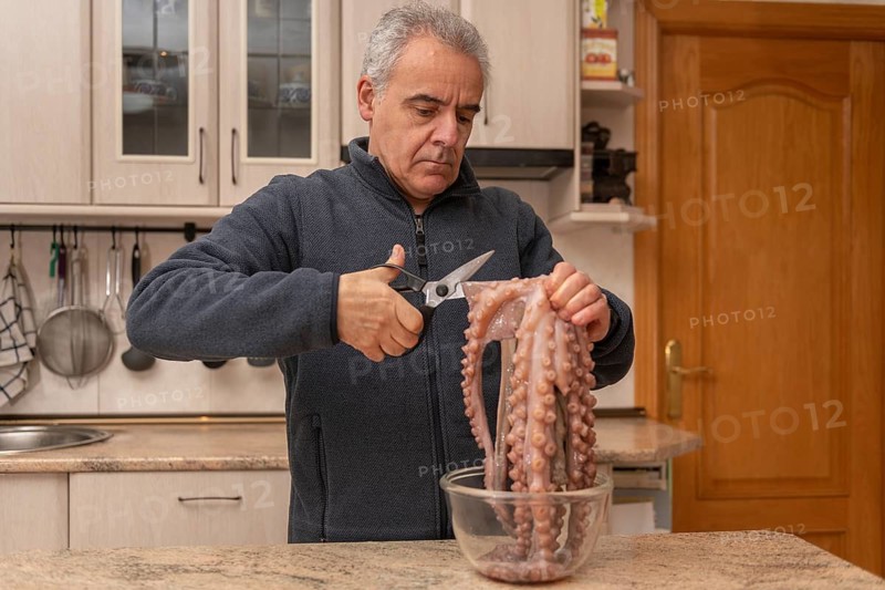 A man in a home kitchen uses scissors to clean a fresh octopus, demonstrating a step in seafood preparation. The kitchen setting is cozy and neatly organized