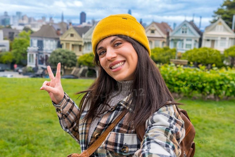 Young woman smiling and holding up two fingers for a peace sign, enjoying her urban vacation with the iconic painted ladies and city skyline in the background