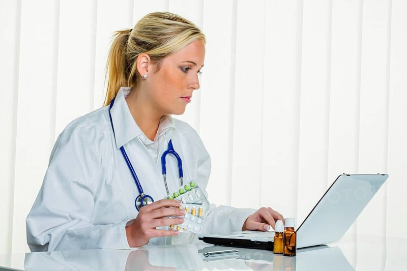 A blonde doctor with a stethoscope prescribing medication, computer, laptop, practice, office studio shot, Austria