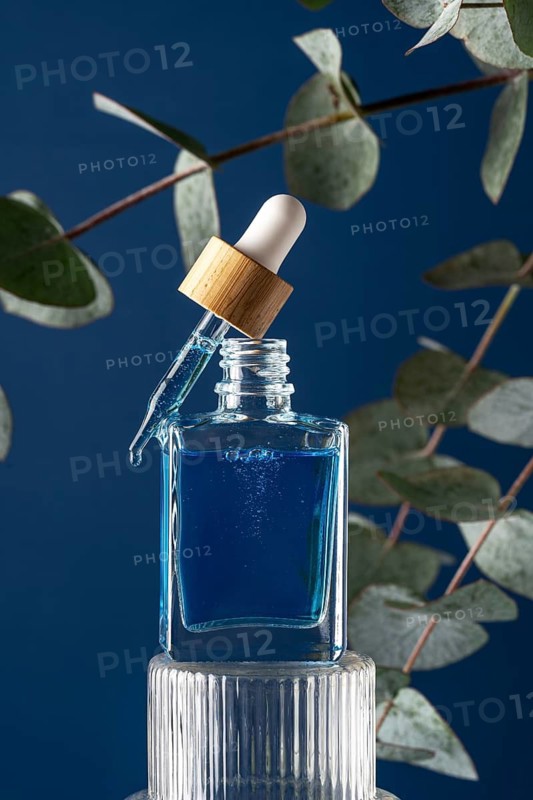 A glass dropper bottle filled with blue cooling face serum or oil designed for men, set against a blue background surrounded by eucalyptus